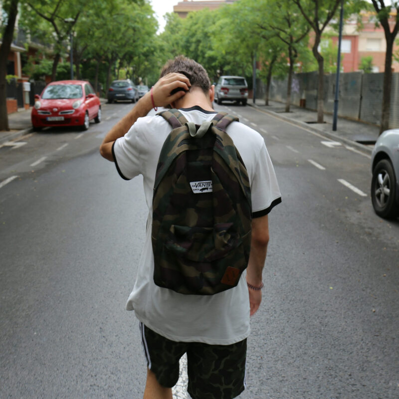 young boy with backpack scratches his head as he walks down street with cars